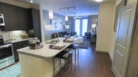 This thoughtfully designed single-floor residence showcases a seamless open-concept layout that connects the kitchen, dining, and living areas into one cohesive, welcoming space. The kitchen is anchored by a granite-topped island with bar seating, offering both functionality and an elegant focal point. Pendant lighting with sleek metallic fixtures hangs above the island, complementing the track lighting and adding a contemporary touch. Dark hardwood-style flooring runs throughout, creating a warm and sophisticated foundation that ties the entire space together. The living area is generously proportioned, featuring a ceiling fan and large sliding glass doors dressed with floor-to-ceiling curtains that invite abundant natural light and lead to a private outdoor area. Soft, neutral wall tones accented by a bold blue feature wall lend depth and visual interest to the interior. The space is further enhanced by stylish furnishings, layered lighting, and curated décor that contribute to a polished, modern ambiance. Overall, this residence offers a refined and functional living environment suited for both everyday comfort and entertaining.