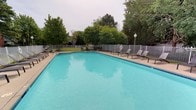 The outdoor pool area offers a generously sized swimming pool with clear turquoise water, making it a central feature of the space. The pool is surrounded by a wide concrete deck that provides ample room for movement and relaxation. Several lounge chairs are arranged along the poolside, offering residents and guests convenient spots to rest and enjoy the outdoors. A white metal perimeter fence encloses the entire area, providing both safety and a sense of seclusion. Mature deciduous and evergreen trees line the background beyond the fence, contributing to a natural, private setting and offering partial shade during warmer months. A classic lamp post stands near one corner of the deck, suggesting the area is also functional during evening hours. The overall ambiance is calm and well-maintained, with a clean, open layout that balances recreational space with comfort. The tree coverage and fencing create a sheltered environment, making it an appealing outdoor amenity suited for warm-weather leisure and recreation.