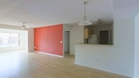 This single-floor space is designed with an open-concept layout that maximizes flow and functionality. The living area makes an immediate visual impact with a striking terracotta-red accent wall that adds warmth and character to the otherwise neutral interior. Light-toned hardwood flooring runs throughout, contributing to a clean, airy aesthetic and visually connecting the spaces.

The kitchen is integrated into the open floor plan, separated from the living area by a raised breakfast bar or peninsula counter, which provides both a casual dining surface and a natural boundary between the spaces. Light wood cabinetry lines the kitchen walls, complementing the overall warm tone of the interior, while a refrigerator and additional appliances are visible within the kitchen zone. A pendant light fixture hangs above the bar area, providing focused task lighting and an element of understated style.

The layout also includes access to additional rooms or storage, suggested by a doorway visible beyond the accent wall. The white ceilings and neutral wall tones throughout help reflect light and give the space an open, uncluttered feel. Overall, this space combines practical design with distinct visual character, making it both livable and inviting.