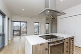 This contemporary single-floor residence showcases a thoughtfully designed open-concept layout with a clean, modern aesthetic throughout. The kitchen is a standout feature, equipped with high-gloss white upper cabinetry, warm wood-toned lower cabinets, a large central island with a white quartz countertop, stainless steel appliances including a French door refrigerator and range, and a striking suspended range hood — all set atop light-toned hardwood flooring. The adjoining living area is bright and expansive, flooded with natural light through floor-to-ceiling black-framed windows that frame sweeping city skyline views, with direct access to an outdoor balcony. Multiple bedrooms feature plush neutral carpeting, ceiling fans, and closets, offering comfortable and versatile spaces. One bedroom benefits from its own access to outdoor views through a large black-framed window. The bathrooms are finished in a consistent modern style, featuring warm wood vanities with white quartz countertops, rectangular undermount sinks, contemporary hardware, large-format tile flooring, and frameless glass shower enclosures. The primary bathroom includes a dual-sink vanity and a walk-in closet. Throughout the space, a neutral white and gray palette reinforces the clean, contemporary character.