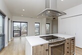 This contemporary single-floor residence showcases a thoughtfully designed open-concept layout with a clean, modern aesthetic throughout. The kitchen is a standout feature, equipped with high-gloss white upper cabinetry, warm wood-toned lower cabinets, a large central island with a white quartz countertop, stainless steel appliances including a French door refrigerator and range, and a striking suspended range hood — all set atop light-toned hardwood flooring. The adjoining living area is bright and expansive, flooded with natural light through floor-to-ceiling black-framed windows that frame sweeping city skyline views, with direct access to an outdoor balcony. Multiple bedrooms feature plush neutral carpeting, ceiling fans, and closets, offering comfortable and versatile spaces. One bedroom benefits from its own access to outdoor views through a large black-framed window. The bathrooms are finished in a consistent modern style, featuring warm wood vanities with white quartz countertops, rectangular undermount sinks, contemporary hardware, large-format tile flooring, and frameless glass shower enclosures. The primary bathroom includes a dual-sink vanity and a walk-in closet. Throughout the space, a neutral white and gray palette reinforces the clean, contemporary character.