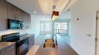 This single-floor residence showcases a contemporary open-concept layout that seamlessly connects the kitchen and living areas. The kitchen is well-equipped with dark wood cabinetry extending to the ceiling for ample storage, stainless steel appliances including a refrigerator and electric range, a built-in microwave, and a central island featuring a double stainless steel undermount sink with a pendant light fixture above. Light wood-tone flooring anchors the kitchen space, transitioning into plush carpeting throughout the living and sleeping areas.

The living area is bright and airy, benefiting from large windows and a sliding glass door that opens to a private balcony, offering views of the surrounding urban streetscape. Natural light flows generously through the space, enhancing its open and welcoming ambiance.

The bedroom is a quiet, carpeted retreat with large windows framing city views and access to the balcony, providing additional natural light and ventilation.

The bathroom features a dark wood vanity with taupe countertops, an undermount sink, a large mirror with overhead lighting, tile flooring, and a walk-in shower, combining functionality with a clean, modern aesthetic throughout.