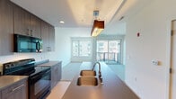 This single-floor residence showcases a contemporary open-concept layout that seamlessly connects the kitchen and living areas. The kitchen is well-equipped with dark wood cabinetry extending to the ceiling for ample storage, stainless steel appliances including a refrigerator and electric range, a built-in microwave, and a central island featuring a double stainless steel undermount sink with a pendant light fixture above. Light wood-tone flooring anchors the kitchen space, transitioning into plush carpeting throughout the living and sleeping areas.

The living area is bright and airy, benefiting from large windows and a sliding glass door that opens to a private balcony, offering views of the surrounding urban streetscape. Natural light flows generously through the space, enhancing its open and welcoming ambiance.

The bedroom is a quiet, carpeted retreat with large windows framing city views and access to the balcony, providing additional natural light and ventilation.

The bathroom features a dark wood vanity with taupe countertops, an undermount sink, a large mirror with overhead lighting, tile flooring, and a walk-in shower, combining functionality with a clean, modern aesthetic throughout.