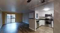 This single-floor space offers a thoughtfully designed open-concept layout that seamlessly connects the living and kitchen areas. The kitchen is well-appointed with crisp white cabinetry, dark countertops, and a full suite of stainless steel appliances, including a dishwasher, microwave, and range. A peninsula with an integrated sink serves as both a functional workspace and a natural divider between the kitchen and the open living area, enhancing the flow of the space. The living area is finished with rich, dark hardwood-style flooring that extends throughout, adding warmth and visual continuity. Crown molding frames the ceiling, lending a polished, finished look to the interior. A ceiling fan with a light fixture keeps the space comfortable and well-lit. Natural light filters in through multiple windows, including a sliding glass door that opens to an outdoor area, creating a bright and airy atmosphere. The neutral gray wall tones complement both the flooring and the kitchen finishes, giving the space a cohesive and modern aesthetic. Overall, the layout and finishes make this a practical and attractive single-floor living space.