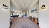 Located in East Wenatchee, WA, this single-story community clubhouse offers a thoughtfully designed gathering space with a welcoming atmosphere throughout. The entrance greets visitors with soaring ceilings, large glass double doors, and an abundance of natural light streaming through oversized windows that frame views of the surrounding outdoor pool area.

The expansive main living area features warm, neutral tones, textured carpeting, and recessed lighting. A tiled fireplace with a wall-mounted TV serves as a focal point, complemented by comfortable leather seating. Adjacent to this lounge area is a functional kitchenette outfitted with wood cabinetry, granite-style countertops, a built-in microwave, and a compact refrigerator — ideal for community events.

A connected hallway leads to a bright, windowed game room equipped with a ping-pong table, bookshelf, and café-style seating, making it a versatile recreational space. Two clean, tile-floored bathrooms serve the facility, one of which is ADA-accessible with grab bars for added convenience.

Throughout the space, large windows and glass doors maintain a strong visual connection to the outdoors, while the open floor plan and high ceilings contribute to an airy, inviting ambiance suitable for a variety of community gatherings.