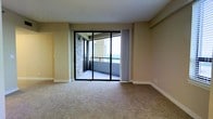 This single-floor space features a bright, open living area finished with wall-to-wall carpet in a neutral beige tone, contributing to a clean and understated aesthetic. The walls are painted in a soft, warm beige, complementing the overall light palette of the interior. A prominent set of black-framed sliding glass doors serves as the focal point of the main living area, providing direct access to a private balcony. The balcony, finished with a light-toned floor surface and enclosed by a metal railing, offers elevated outdoor views of the surrounding landscape, with glimpses of greenery visible in the distance. Sheer white curtains flank the sliding doors, allowing diffused natural light to filter into the living space throughout the day. The elevated position of the unit enhances the sense of openness and airiness within the interior. Additional architectural details include clean-lined door frames and standard baseboard trim throughout. A doorway visible along one wall suggests access to additional interior rooms. The overall ambiance is calm and functional, well-suited to a variety of living arrangements.