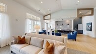 Located at 500 River Knoll Way in Columbus, Georgia, this single-story space offers a generously sized, open-plan layout combining a kitchen and living area under one cohesive design. The architecture is defined by soaring vaulted ceilings that amplify the sense of volume throughout, complemented by recessed lighting and large windows that flood the interior with natural light and offer views of the surrounding outdoor area, including a pool.

The living area features a bold teal accent wall with built-in shelving and an inset television, paired with light hardwood-style flooring and neutral carpeting. Seating areas are thoughtfully arranged to encourage conversation and comfort, while a gallery wall displaying large-scale abstract artwork adds a sophisticated visual anchor near the entryway.

The kitchen is a standout feature, showcasing a substantial white quartz island with bar seating, matte grey cabinetry, and a striking full-height grey brick tile accent wall. Three oversized black pendant lights hang above the island, providing both task lighting and a bold design statement. A wet bar area with under-counter appliances and additional counter space enhances the kitchen's functionality. The seamless flow between the kitchen and living zones makes this space well-suited for both everyday use and entertaining.