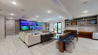 Located in Winter Park, Florida, this well-appointed community club room offers a sophisticated and versatile gathering space spread across a single floor. The interior design blends modern and transitional styles, with light-toned tile flooring, tray ceilings with recessed lighting, and textured stacked-stone accent walls that add warmth and visual interest throughout.

The main lounge area serves as the social hub, featuring a large sectional sofa arrangement anchored by a bold multi-screen media wall displaying vibrant artwork and nature imagery. A long communal bar-height table with upholstered ottool seating occupies the center of the space, complemented by built-in shelving units ideal for décor and personal items. A wet bar with wood cabinetry and stainless steel fixtures provides an elegant service area, and large windows and glass doors invite abundant natural light and views of the surrounding greenery.

A separate dining zone includes upholstered booth seating along a stone-clad accent wall, with wall-mounted screens for additional entertainment. Adjacent to the main area, a private conference room features a geometric accent wall, a long conference table with high-back chairs, a wall-mounted display, and striking geometric pendant lighting, making it well-suited for meetings or focused work sessions. Curated abstract artwork is displayed throughout, elevating the overall ambiance of this thoughtfully designed community space.
