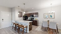 This single-floor residence offers a thoughtfully designed layout that balances comfort and functionality throughout. The open-concept kitchen is a standout feature, showcasing rich dark-brown cabinetry, light granite countertops, and black appliances including a refrigerator, electric range, microwave, and dishwasher. A peninsula with bar seating and stylish pendant lighting serves as both a prep space and a casual gathering spot, flowing seamlessly into the dining area. The dining room is an intimate yet elegant space, highlighted by a chandelier overhead and light hardwood-style flooring that continues throughout the main living areas. The living room offers a welcoming atmosphere with a ceiling fan, natural light from large windows, and direct access to an entry door. The bedroom is a comfortable retreat featuring plush carpet, a ceiling fan, and a generously sized walk-in closet with wire shelving for ample storage. The full bathroom is finished with granite countertops, dark cabinetry, a large mirror, and a shower-tub combination. Completing the unit is a dedicated in-unit laundry space equipped with a full-size washer and dryer and additional wire shelving above for extra storage convenience.