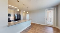 This single-floor residence is designed with a contemporary open-concept layout that seamlessly connects the kitchen and living areas. The kitchen serves as a central focal point, anchored by a large island finished with speckled granite countertops, an undermount sink with a pull-down faucet, and a built-in dishwasher. Gray shaker-style cabinetry with dark hardware pairs with black appliances, including a refrigerator and range, while three cylindrical pendant lights suspended above the island provide focused task lighting. Hardwood-style flooring flows throughout the main living and kitchen areas, adding warmth and continuity.

A hallway off the main living space features built-in open shelving on one side, offering practical storage and display options, and leads toward the bedroom and bathroom areas. The bathroom is appointed with a granite-topped vanity, gray cabinetry matching the kitchen aesthetic, a large mirror, and a shower with tiled surround. The bedroom is carpeted for comfort and benefits from a large window that allows generous natural light and outdoor views, complemented by a ceiling fan. Neutral gray walls and clean white trim throughout give the space a cohesive, modern feel.
