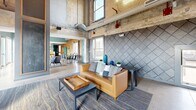 Located at 1375 Willow St in Minneapolis, MN, this thoughtfully designed single-floor communal space masterfully merges industrial heritage with contemporary style. Exposed concrete walls, raw structural columns, and soaring double-height ceilings give the main lounge area a dramatic, loft-like character, while a tiled fireplace with a warm wood mantel and flat-screen TV serves as a striking focal point. Patterned accent walls, layered area rugs, and curated furnishings — including a caramel leather sofa and plush seating — create an inviting atmosphere throughout.

The dining area is anchored by a substantial solid wood conference-style table surrounded by modern chairs, set against a full wall of richly textured wood paneling and large windows that frame views of the surrounding Minneapolis skyline. Exposed ductwork and industrial-style HVAC elements add an authentic urban edge.

The kitchen is outfitted with warm wood cabinetry, quartz countertops, a mosaic tile backsplash, floating wood shelves, and pendant lighting, with polished concrete floors tying the space together. A set of expansive floor-to-ceiling sliding glass doors opens directly onto a spacious outdoor terrace, flooding the interior with natural light and providing seamless access to open-air city views — a standout feature of this vibrant, multifunctional space.