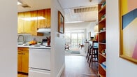 This single-floor residence showcases a cohesive mid-century modern aesthetic carried throughout every room. The kitchen is outfitted with warm wood cabinetry, white appliances including a retro-style Frigidaire refrigerator, tile backsplash, and durable wood-look flooring — a charming nod to classic design. A built-in open shelving unit transitions the kitchen area into a dining space with bar seating, keeping the layout connected and functional.

The living room is a standout, flooded with natural light through expansive floor-to-ceiling windows that open to a private balcony overlooking a leafy outdoor view. Neutral walls and carpet provide a calm backdrop for bold, colorful furnishings and a large statement area rug. Built-in wood shelving adds both character and practical display space.

The bedroom continues the retro-inspired theme with plush carpeting, a reach-in closet with louvered doors offering generous storage, and a natural window that brings in soft light. Accent wallpaper and open shelving lend personality to the space.

Throughout, the residence blends warm wood tones, vibrant accents, and clean-lined furniture to create an energetic yet comfortable atmosphere true to mid-century modern design.
