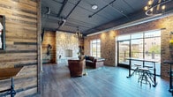 The Polo Lounge in Lemont, Illinois is a thoughtfully designed single-floor venue that masterfully blends rustic charm with industrial character. The open-plan layout offers a generous, comfortable footprint ideal for socializing and entertaining.

The space is anchored by a striking floor-to-ceiling natural stone fireplace, which serves as a dramatic focal point in the lounge area. Rich leather Chesterfield sofas and tufted armchairs surround the hearth, creating an inviting and sophisticated seating arrangement atop a hardwood floor in a cool, weathered grey tone.

Exposed brick walls run throughout the space, complemented by reclaimed wood plank accent walls that add warmth and texture. The industrial aesthetic is reinforced by an exposed ceiling featuring dark painted ductwork, conduit, and Edison-bulb chandelier lighting that casts a warm, ambient glow.

A built-in bar area with light cabinetry and a white quartz countertop provides a sleek contrast to the rustic surroundings. Nearby, high-top tables with industrial-style stools offer casual seating near a wall-mounted television. Polo-themed artwork and memorabilia add a distinctive character to the space.

Large floor-to-ceiling windows and glass doors open to an outdoor balcony, flooding the interior with natural light and offering scenic views of the surrounding Lemont streetscape and waterway, seamlessly connecting the indoors with the outdoors.