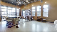 This 770 square foot single-floor space in Salt Lake City, UT, showcases a striking blend of industrial and modern design elements. The interior is characterized by high ceilings with exposed ductwork and conduits, creating an open, loft-like atmosphere. Large windows allow ample natural light to flood the space, enhancing its airy feel.

The area is divided into multiple functional zones. A contemporary kitchenette features sleek blue cabinetry, stainless steel appliances, and a unique curved island with bar seating. Adjacent to this is a versatile seating area with a mix of chairs and tables, ideal for casual meetings or work sessions.

The lounge areas are thoughtfully designed with comfort and style in mind. One section boasts plush, modern seating arrangements including cream-colored pod chairs and a striped sofa, complemented by a striking black marble coffee table. A large black and white landscape photograph serves as a focal point, adding depth to the space.

The floor throughout is polished concrete, consistent with the industrial aesthetic while providing a neutral backdrop for the eclectic furniture. Lighting is a key feature, with a combination of pendant lights, wall sconces, and recessed lighting creating a warm, inviting ambiance.

Overall, this space offers a flexible, contemporary environment suitable for various purposes, from creative workspaces to trendy social gatherings.