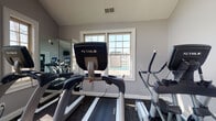 Located at 2223 Craigside Dr in Columbus, Ohio, this single-floor fitness center is purpose-built to support an active lifestyle. The space is equipped with professional-grade TRUE Brand cardio machines, including treadmills, ellipticals, and stationary bikes, arranged to maximize usability and flow throughout the room. Large, grid-pane windows allow ample natural light to fill the space, creating a bright and energizing workout environment with views to the outdoors. Full-length wall mirrors line one side of the room, visually expanding the space while allowing users to monitor their form during exercise. The neutral gray walls and dark patterned carpet flooring provide a clean, focused aesthetic well-suited to a fitness setting. Overhead ventilation ensures proper air circulation throughout the room. The overall design is functional and well-appointed, offering residents or users a dedicated, well-lit space for cardiovascular training in a comfortable and organized setting.