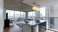 Located at 3737 Chestnut Street in Philadelphia's University City neighborhood, this contemporary single-floor space showcases a refined, modern aesthetic that seamlessly blends form and function. The open-concept layout connects the kitchen and living area in a fluid, cohesive design, creating a sense of expansive, uninterrupted space.

The kitchen is a standout feature, centered around a large island with polished granite countertops, an undermount sink, and dark cabinetry with clean, handleless lines. A sleek pendant light fixture hangs above the island, providing both task lighting and a stylish focal point. Stainless steel appliances, including a dishwasher integrated into the island, complete the sophisticated culinary space.

The living area flows naturally from the kitchen, anchored by a contemporary seating arrangement atop a textured area rug over warm hardwood flooring. Exposed concrete ceilings lend an industrial character to the space, contrasting beautifully with the refined finishes throughout.

Perhaps the most dramatic feature is the wraparound floor-to-ceiling windows that flood the interior with abundant natural light while framing sweeping views of the Philadelphia skyline. The building's urban surroundings are fully on display, making the cityscape an ever-present backdrop to daily life in this architecturally compelling space.