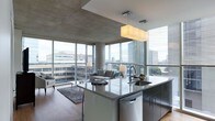 Located at 3737 Chestnut Street in Philadelphia's University City neighborhood, this contemporary single-floor space showcases a refined, modern aesthetic that seamlessly blends form and function. The open-concept layout connects the kitchen and living area in a fluid, cohesive design, creating a sense of expansive, uninterrupted space.

The kitchen is a standout feature, centered around a large island with polished granite countertops, an undermount sink, and dark cabinetry with clean, handleless lines. A sleek pendant light fixture hangs above the island, providing both task lighting and a stylish focal point. Stainless steel appliances, including a dishwasher integrated into the island, complete the sophisticated culinary space.

The living area flows naturally from the kitchen, anchored by a contemporary seating arrangement atop a textured area rug over warm hardwood flooring. Exposed concrete ceilings lend an industrial character to the space, contrasting beautifully with the refined finishes throughout.

Perhaps the most dramatic feature is the wraparound floor-to-ceiling windows that flood the interior with abundant natural light while framing sweeping views of the Philadelphia skyline. The building's urban surroundings are fully on display, making the cityscape an ever-present backdrop to daily life in this architecturally compelling space.