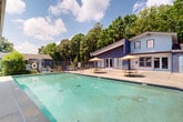 Located at 600 Antlers Lane in Charlotte, NC, this outdoor amenity space centers around a generously sized swimming pool set within a well-maintained, fenced pool deck. The pool area is surrounded by a broad concrete surround that provides ample space for lounging and socializing. Multiple seating configurations are distributed throughout the deck, including cushioned lounge chairs ideal for sunbathing and a covered pergola-style structure on one side offering shaded relief. Umbrella-shaded bistro tables and chairs near the main building provide additional spots for relaxation and casual gathering.

The pool area is enclosed by a wrought iron fence, ensuring security and privacy while maintaining open sightlines to the mature trees that frame the perimeter. This lush, green backdrop of tall pines and deciduous trees contributes to a tranquil, retreat-like atmosphere. An outdoor lounge seating arrangement with cushioned chairs and a sofa is positioned along the building's exterior, creating an inviting social zone.

The adjacent building, finished in light blue and navy tones, features large windows and glass-paned doors that open directly onto the pool deck, reinforcing the indoor-outdoor connection. The single-level layout of the surrounding structures ensures easy, accessible flow throughout the entire outdoor space, making it a functional and attractive communal amenity.