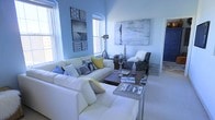 This single-floor space centers around a thoughtfully designed living area that combines comfort with contemporary style. The room is defined by its striking powder-blue walls, which create a calm, airy atmosphere throughout. Natural light streams in through multiple windows, including an arched window that adds an architectural detail to the space, brightening the room and enhancing its open feel.

The living area features a generous white sectional sofa adorned with an eclectic mix of decorative pillows in blue, grey, and yellow tones, adding visual interest and warmth. A coordinating dark-upholstered accent chair with a plush white throw provides an additional seating option. Two coffee tables anchor the seating arrangement, offering both functionality and style.

Wall art is a prominent feature, with a large abstract canvas in cool grey and white tones displayed on one wall, while a curated grid of black-and-white prints and patterned artwork decorates another. A vintage-style tripod floor lamp adds an industrial accent. A bold cobalt-blue door visible in the background introduces a vibrant pop of color, tying the room's palette together with personality and intention.