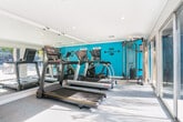 Located at 530 Sunnyview Dr in Pinole, California, this fitness center offers a well-equipped workout space across a single floor. The gym's interior is defined by a clean, modern aesthetic, with crisp white ceilings accented by recessed lighting and a bold teal accent wall that adds a vibrant, energizing atmosphere. Full-length mirrored walls line one side of the room, visually expanding the space and supporting proper form during exercise. The main workout area is outfitted with multiple treadmills and a cable resistance machine, providing a solid range of cardio and strength training options. Motivational wall decals reinforce the gym's active atmosphere. Large sliding glass doors span the length of one wall, flooding the space with natural light and offering direct views and access to an outdoor pool deck, complete with seating and mature palm trees. This seamless indoor-outdoor connection enhances the overall workout experience. A bathroom is conveniently located within the facility, accompanied by towel bars stocked with dark towels for guest use. The entrance features a distinctive teal door, tying together the space's color palette. Durable, light-toned carpet flooring completes the practical and welcoming design of this community fitness amenity.