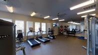 The fitness center is a thoughtfully designed, single-floor space that offers a comprehensive range of equipment for a well-rounded workout. The room is illuminated by a combination of recessed lighting and overhead fluorescent fixtures, supplemented by natural light streaming in through multiple windows fitted with blinds along one wall. A ceiling fan provides additional air circulation, contributing to a comfortable exercise environment.

The cardio section is prominently featured, housing multiple treadmills and an elliptical machine positioned along the windowed wall, allowing users to enjoy natural light during their workouts. A wall-mounted flat-screen television is installed nearby, providing entertainment options for those on cardio equipment.

On the opposite side of the room, a large mirror runs along the wall, visually expanding the space and allowing for form checks during exercise. A well-stocked dumbbell rack offers a range of free weights, complemented by a selection of medicine balls and stability balls for functional training. The flooring throughout is a durable, speckled rubber surface, providing both safety and comfort underfoot. Overall, the space presents a clean, functional, and inviting atmosphere for fitness enthusiasts.