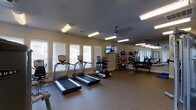The fitness center is a thoughtfully designed, single-floor space that offers a comprehensive range of equipment for a well-rounded workout. The room is illuminated by a combination of recessed lighting and overhead fluorescent fixtures, supplemented by natural light streaming in through multiple windows fitted with blinds along one wall. A ceiling fan provides additional air circulation, contributing to a comfortable exercise environment.

The cardio section is prominently featured, housing multiple treadmills and an elliptical machine positioned along the windowed wall, allowing users to enjoy natural light during their workouts. A wall-mounted flat-screen television is installed nearby, providing entertainment options for those on cardio equipment.

On the opposite side of the room, a large mirror runs along the wall, visually expanding the space and allowing for form checks during exercise. A well-stocked dumbbell rack offers a range of free weights, complemented by a selection of medicine balls and stability balls for functional training. The flooring throughout is a durable, speckled rubber surface, providing both safety and comfort underfoot. Overall, the space presents a clean, functional, and inviting atmosphere for fitness enthusiasts.