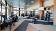 This fitness center offers a thoughtfully designed, single-floor workout facility that balances functionality with contemporary style. The space is defined by its striking embossed tin ceiling, which adds a distinctive architectural character, complemented by recessed lighting that ensures the entire area is well-lit throughout. Large floor-to-ceiling windows line the perimeter, flooding the interior with abundant natural light and providing open views of the surrounding streetscape.

The flooring features a bold geometric pattern of alternating light and dark tones, creating visual zones that naturally separate the cardio and strength training areas. The cardio section is outfitted with a comprehensive selection of equipment, including elliptical trainers, stationary bikes, and treadmills, all positioned to take advantage of the expansive window views.

The strength training zone is equally well-appointed, featuring a multi-station cable machine, a full dumbbell rack with a wide range of weights, and multiple adjustable benches. Full-length mirrors line the walls of this section, enhancing the sense of space and allowing users to monitor their form.

A dedicated functional training corner provides stability balls, medicine balls, resistance bands, and exercise mats, catering to a variety of workout styles. Warm orange accent walls and brick detailing add a vibrant, energetic ambiance throughout the space.