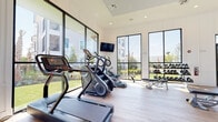 This spacious fitness center is designed with both functionality and aesthetics in mind, offering a bright and motivating environment for a comprehensive workout experience. The space is defined by its striking floor-to-ceiling windows with sleek black frames, which line multiple walls and flood the interior with natural light while providing unobstructed views of the surrounding outdoor greenery, pool area, and palm trees.

The light wood flooring runs throughout, complementing the clean white walls and recessed lighting overhead. A ceiling fan is integrated into a raised tray ceiling detail in one section of the room, enhancing airflow and comfort during workouts.

The fitness center is comprehensively equipped across multiple zones. The cardio area features treadmills, stair climbers, ellipticals, a stationary bike, and a rowing machine. The strength training section includes a full rack of dumbbells, adjustable benches, cable machines, and selectorized weight machines suitable for targeting all major muscle groups. A dedicated functional training zone offers medicine balls, battle ropes, and additional free-motion equipment. Wall-mounted televisions are positioned throughout for entertainment. Full-length mirrors further enhance the sense of openness while providing visual feedback during training sessions.