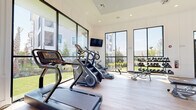 This spacious fitness center is designed with both functionality and aesthetics in mind, offering a bright and motivating environment for a comprehensive workout experience. The space is defined by its striking floor-to-ceiling windows with sleek black frames, which line multiple walls and flood the interior with natural light while providing unobstructed views of the surrounding outdoor greenery, pool area, and palm trees.

The light wood flooring runs throughout, complementing the clean white walls and recessed lighting overhead. A ceiling fan is integrated into a raised tray ceiling detail in one section of the room, enhancing airflow and comfort during workouts.

The fitness center is comprehensively equipped across multiple zones. The cardio area features treadmills, stair climbers, ellipticals, a stationary bike, and a rowing machine. The strength training section includes a full rack of dumbbells, adjustable benches, cable machines, and selectorized weight machines suitable for targeting all major muscle groups. A dedicated functional training zone offers medicine balls, battle ropes, and additional free-motion equipment. Wall-mounted televisions are positioned throughout for entertainment. Full-length mirrors further enhance the sense of openness while providing visual feedback during training sessions.