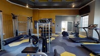 Located at 9874 E Arizona Dr in Aurora, Colorado, this well-appointed fitness center offers a comprehensive workout environment across a single level. The space is immediately distinguished by its bold design palette — vibrant mustard yellow accent walls contrast with cool gray tones, while the eye-catching hexagonal patterned flooring in yellow and gray reinforces the energetic aesthetic throughout. Tray ceilings with pendant lighting add an elevated architectural detail rarely found in fitness facilities.

The main workout area is generously equipped with a full range of cardio machines, including multiple treadmills, ellipticals, and a rowing machine, all arranged to allow comfortable spacing between users. A dedicated strength training zone features cable machines, a multi-use weight station, adjustable benches, and a dumbbell rack, providing options for a complete resistance workout.

A separate studio room offers a more intimate space for focused training, complete with a spin bike, step platforms, kettlebells, a ballet barre, wall-mounted mirrors, and a large flat-screen TV. Motivational text adorns the yellow accent wall, enhancing the room's energizing atmosphere.

Large windows throughout the facility invite ample natural light, and glass doors connect the fitness center to an adjacent amenity space, creating a bright and open feel across the entire facility.