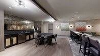 Located at 3444 S Emerson St in Englewood, Colorado, this single-floor amenity lounge is a well-appointed communal space designed for socializing and entertainment. The layout is thoughtfully divided into distinct activity zones, all unified by a cohesive modern aesthetic featuring textured geometric wallcoverings, warm wood-look flooring, and exposed dark ceiling beams that add architectural character.

The central gathering area is anchored by a dedicated poker and game table surrounded by cushioned chairs, making it ideal for game nights and social events. Adjacent to it, a sleek wet bar runs along one wall, complete with dark countertops, cane-front cabinetry, a stainless beverage refrigerator, and an antiqued mirror backsplash that adds depth and glamour to the space. A high-top bar counter with bar-height seating provides an additional area for casual dining or drinks.

A separate lounge nook offers a more relaxed setting, furnished with a generous L-shaped sectional sofa in deep green velvet, a round dark coffee table, and layered area rugs over plush carpeting. Contemporary framed artwork is displayed throughout, complemented by stylish wall sconces that provide warm, ambient lighting. The overall ambiance is sophisticated yet inviting, reflecting a modern club-like atmosphere suited for both lively gatherings and quiet relaxation.