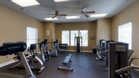 This fitness center offers a comprehensive workout environment designed to accommodate a range of exercise routines. The space is finished with durable rubber flooring throughout, providing a safe and functional surface for all types of training activities. Warm-toned walls create an inviting atmosphere, while multiple large windows fitted with white blinds allow generous natural light to fill the room, enhancing the overall sense of openness. Two ceiling fans with integrated lighting fixtures ensure comfortable air circulation during workouts. The cardio section features elliptical trainers and step machines, ideal for sustained aerobic exercise. A dedicated free weights area includes a full dumbbell rack, accompanied by a flat workout bench for versatile strength training exercises. On the opposite side of the room, a cable and pulley machine provides options for targeted resistance training, alongside additional selectorized weight equipment. Informational signage is posted on the wall, offering guidance for proper equipment use. The layout is thoughtfully arranged to provide ample space between machines, allowing for comfortable movement and safe exercise throughout the facility.
