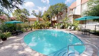 Located at 2200 S Gessner Rd in Houston, TX, this single-floor residential unit offers a comfortable and well-appointed living experience in a lushly landscaped community setting. The bedroom is a standout feature, showcasing warm hardwood flooring, crown molding, a ceiling fan, and a tray ceiling that adds architectural interest. Large sliding glass doors flood the room with natural light and open directly to a private outdoor patio, blending indoor and outdoor living seamlessly. A dedicated closet with built-in shelving and hanging rods provides ample storage. The hallway connects to a tiled utility area housing a stacked washer and dryer unit, offering the convenience of in-unit laundry. The interior carries a neutral, warm color palette throughout, creating a calm and cohesive ambiance. Beyond the unit itself, residents enjoy access to a beautifully maintained outdoor pool area — a generously sized, oval-shaped pool with clear turquoise water, surrounded by a tiled deck, lounge seating, and abundant tropical greenery including palms and mature shade trees, all enclosed within an iron fence. The overall atmosphere is tranquil and resort-like, making it an inviting place to call home.
