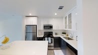 This single-floor space showcases a thoughtfully designed kitchen as its centerpiece. The kitchen features a striking two-tone cabinet scheme, pairing high-gloss white upper cabinets with dark lower cabinets to create a bold, modern contrast. Upper cabinets extend toward the ceiling, maximizing vertical storage, while select glass-front panels add visual interest and an open feel. The countertops are finished in a clean white surface that complements the overall color palette.

Stainless steel appliances, including a full-size refrigerator, electric range with oven, over-the-range microwave, and dishwasher, are seamlessly integrated into the design. A stainless steel undermount sink with a modern arc-neck faucet is positioned along the countertop run, adding both functionality and style. Recessed lighting in the white ceiling provides bright, even illumination throughout. A kitchen island with matching white countertop offers additional workspace and casual seating potential. The polished concrete-style flooring ties the space together, reinforcing the clean, contemporary aesthetic throughout.