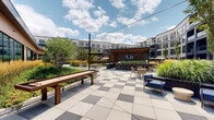 Located at 253 NJ-18 in East Brunswick, New Jersey, this contemporary residential community showcases a striking multi-story building with a sleek architectural design defined by large floor-to-ceiling windows, private balconies, and a refined façade finished in neutral grey tones with bold dark accents. The building's modern exterior is complemented by thoughtfully curated landscaping featuring ornamental grasses, flowering shrubs, and manicured plantings that line the perimeter walkways.

At the heart of the community lies an expansive resort-style outdoor amenity deck. A generously sized swimming pool serves as the centerpiece, surrounded by a wide pool deck furnished with rows of lounge chairs and shade umbrellas, ideal for relaxation and recreation. The pool area is nestled within the building's courtyard, creating a sense of privacy and enclosure while remaining open to the sky.

Beyond the pool, the amenity grounds extend to include multiple distinct outdoor social zones. A central pavilion-style clubhouse structure anchors one section of the deck, featuring warm wood accents and large glass entries that invite natural light. Paved plaza areas rendered in a geometric two-tone tile pattern provide space for seating and gathering, interspersed with raised garden beds, seasonal plantings, and shade trees. The overall ambiance is polished, resort-inspired, and designed for active outdoor living.