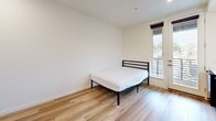 Located at 4746 20th Ave NE in Seattle's University District, this efficiently designed one-bedroom, one-bathroom unit on the fourth floor makes smart use of its intimate footprint. Light hardwood-style flooring runs continuously throughout, lending warmth and visual cohesion to the entire space.

The main living area doubles as the bedroom, featuring a sleek black metal bed frame and a built-in kitchenette along one wall. The kitchenette is outfitted with warm wood-toned cabinetry with matte black hardware, a countertop sink with a modern faucet, a compact refrigerator, and a microwave — all anchored by a pendant light overhead. A set of glass-paneled doors opens directly to a private balcony, filling the room with natural light and offering views of the surrounding Seattle neighborhood.

The bedroom area is generous in proportion relative to the overall unit, with crisp white walls, recessed lighting, and a built-in desk and chair that create a functional workspace. The bathroom is clean and contemporary, featuring a wood-grain vanity with a white countertop, matte black hardware, and a window that brings in additional natural light.

The overall aesthetic is modern and minimal, with a consistent palette of white walls, natural wood tones, and black accents that give the space a cohesive, well-designed feel.