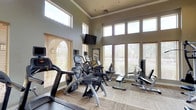 This fitness center is designed to deliver a comprehensive workout experience within a bright, welcoming environment. The space features soaring ceilings accentuated by a row of clerestory windows positioned near the roofline, which work in tandem with large floor-to-ceiling windows along the walls to bathe the interior in abundant natural light. The windows are fitted with adjustable horizontal blinds, offering control over glare while maintaining an airy atmosphere. Neutral-toned walls and patterned carpet flooring contribute to a clean, professional aesthetic throughout.

The facility is outfitted with a strong selection of professional-grade Cybex and comparable cardio equipment, including treadmills, elliptical trainers, a recumbent bike, and an upright cycle. Strength and resistance training needs are addressed with a cable pulley machine and a dedicated weight bench, providing a well-rounded range of exercise options. A wall-mounted flat-screen television is positioned for easy viewing from the cardio machines, adding an entertainment element to workouts. The layout is thoughtfully arranged to allow comfortable movement between stations. Crown molding details add a refined architectural touch, elevating the overall presentation of this functional and inviting fitness space.