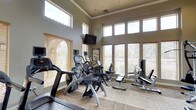 This fitness center is designed to deliver a comprehensive workout experience within a bright, welcoming environment. The space features soaring ceilings accentuated by a row of clerestory windows positioned near the roofline, which work in tandem with large floor-to-ceiling windows along the walls to bathe the interior in abundant natural light. The windows are fitted with adjustable horizontal blinds, offering control over glare while maintaining an airy atmosphere. Neutral-toned walls and patterned carpet flooring contribute to a clean, professional aesthetic throughout.

The facility is outfitted with a strong selection of professional-grade Cybex and comparable cardio equipment, including treadmills, elliptical trainers, a recumbent bike, and an upright cycle. Strength and resistance training needs are addressed with a cable pulley machine and a dedicated weight bench, providing a well-rounded range of exercise options. A wall-mounted flat-screen television is positioned for easy viewing from the cardio machines, adding an entertainment element to workouts. The layout is thoughtfully arranged to allow comfortable movement between stations. Crown molding details add a refined architectural touch, elevating the overall presentation of this functional and inviting fitness space.
