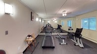 This dedicated fitness space is thoughtfully designed to support a comprehensive workout routine, all contained within a single-floor layout. The room features durable rubber flooring throughout, providing a safe and functional surface for exercise equipment and free movement alike. A full wall of floor-to-ceiling mirrors enhances the sense of space while allowing users to monitor their form during workouts.

The room is equipped with a range of cardio and strength-training equipment, including a treadmill, a stationary upright bike, an elliptical machine, and a flat weight bench with free weights. Ceiling fans provide overhead air circulation, while square wall-mounted sconces offer warm, ambient lighting throughout the space. Additional natural light filters in through multiple windows fitted with horizontal blinds, keeping the room bright and ventilated.

The walls are finished in a neutral cream tone, contributing to a clean and focused atmosphere. Practical touches such as a small side table, folding chairs, and a waste bin add functional convenience to the space. A textured ceiling and crown molding detail the upper perimeter of the room, lending a polished finishing touch. Overall, this fitness room is a practical and well-appointed space suited for regular personal use.