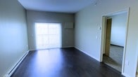 This single-floor residential space offers a clean, open layout designed for comfortable everyday living. The main living area is a standout feature, showcasing rich, dark hardwood flooring that adds warmth and contrast against the neutral, light-gray walls. A large sliding glass door dressed with vertical blinds serves as the room's focal point, allowing abundant natural light to pour in while providing direct access to an outdoor area. The open wall space offers flexibility for a variety of furniture arrangements, making the living area highly adaptable. A wall-mounted unit is positioned above the sliding door, adding a functional element to the space. White baseboards provide a crisp finish throughout, complementing the overall clean aesthetic. A doorway visible to the right leads further into the unit, suggesting additional rooms beyond the main living space. The combination of hardwood flooring, neutral tones, and generous natural light creates a bright, welcoming ambiance. The single-floor layout ensures convenient accessibility throughout, making this a practical and well-appointed living space suited to a range of lifestyles.