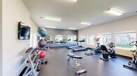 This well-equipped fitness center at 7410 S Hayford Rd in Spokane, Washington offers a comprehensive workout facility within a single, open-plan floor. The space features durable speckled rubber flooring throughout, providing both safety and resilience for a wide range of exercises. Bright flush-mount fluorescent lighting runs across the ceiling, complemented by natural light streaming in through multiple windows that offer views of the surrounding community. The cardio section is outfitted with several treadmills, an elliptical trainer, and a stationary bike, arranged to maximize the available floor space. A dedicated strength training area includes a multi-station cable machine, an adjustable weight bench, a full dumbbell rack, and an assortment of medicine balls and exercise balls on a dedicated storage rack. Resistance bands and additional accessories are neatly organized along the wall. A row of large wall-mounted mirrors along one accent wall visually expands the room and allows users to monitor their form during workouts. A wall-mounted flat-screen television provides entertainment options. Practical amenities include a dual-basin drinking fountain with a white tile surround, interior access doors, and a clearly marked exit. The clean, neutral color palette of white and gray walls creates a bright, motivating atmosphere suited for daily fitness routines.