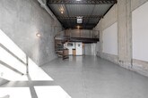Located in the heart of Los Angeles, this two-floor industrial loft at 201 N Westmoreland delivers a raw, urban aesthetic defined by soaring ceilings, exposed steel roof trusses, and corrugated metal roofing. The expansive main floor serves as an open-plan kitchen and living area, characterized by polished concrete floors, bare concrete block walls, and an abundance of natural light streaming through large glass entry doors and a skylight above. A dramatic spiral staircase with warm wood treads and a metal railing anchors the main floor and leads to the upper mezzanine loft — a secondary space framed by a sleek cable railing system and overlooking the ground floor below. The mezzanine benefits from continued exposure to the structural ceiling elements, maintaining the industrial character throughout. Two bathrooms serve the space, one of which features exposed overhead plumbing and piping consistent with the loft's utilitarian design language. Glass block partitioning adds an architectural detail near the kitchen area. The main floor opens to an outdoor area through full-height glass doors, allowing natural light to flood the interior and create striking geometric shadow patterns across the concrete walls. This space offers a distinctive, versatile environment well-suited to live-work or creative studio use.