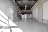 Located in the heart of Los Angeles, this two-floor industrial loft at 201 N Westmoreland delivers a raw, urban aesthetic defined by soaring ceilings, exposed steel roof trusses, and corrugated metal roofing. The expansive main floor serves as an open-plan kitchen and living area, characterized by polished concrete floors, bare concrete block walls, and an abundance of natural light streaming through large glass entry doors and a skylight above. A dramatic spiral staircase with warm wood treads and a metal railing anchors the main floor and leads to the upper mezzanine loft — a secondary space framed by a sleek cable railing system and overlooking the ground floor below. The mezzanine benefits from continued exposure to the structural ceiling elements, maintaining the industrial character throughout. Two bathrooms serve the space, one of which features exposed overhead plumbing and piping consistent with the loft's utilitarian design language. Glass block partitioning adds an architectural detail near the kitchen area. The main floor opens to an outdoor area through full-height glass doors, allowing natural light to flood the interior and create striking geometric shadow patterns across the concrete walls. This space offers a distinctive, versatile environment well-suited to live-work or creative studio use.