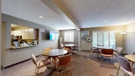 Located at 7700 Cahill Rd in Edina, Minnesota, this single-floor community space is thoughtfully designed to accommodate gatherings, meetings, and everyday social activity. The main lounge area serves as the heart of the space, featuring multiple round tables with chairs arranged across a warm, carpeted floor. A wall-mounted flat-screen TV, plantation-shuttered windows allowing ample natural light, and a wall-unit air conditioner contribute to a comfortable, functional atmosphere. Decorative wall art, including a sculptural tree piece and floral accents, adds a welcoming character to the room. Adjacent to the main lounge, a compact kitchenette is outfitted with stainless steel appliances — including a full-size refrigerator, electric range, and over-the-range microwave — along with generous upper and lower cabinetry and a counter-height pass-through that connects to the main area. A dedicated private meeting room offers a quieter setting with its own round table, seating, decorative wall art, and a floor lamp. A storage area with overhead shelving, hanging rods, and space for folding chairs and equipment rounds out the layout, alongside a full bathroom. The space flows efficiently, making it well-suited for community events, resident gatherings, or small group meetings.