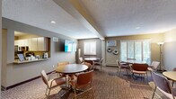 Located at 7700 Cahill Rd in Edina, Minnesota, this single-floor community space is thoughtfully designed to accommodate gatherings, meetings, and everyday social activity. The main lounge area serves as the heart of the space, featuring multiple round tables with chairs arranged across a warm, carpeted floor. A wall-mounted flat-screen TV, plantation-shuttered windows allowing ample natural light, and a wall-unit air conditioner contribute to a comfortable, functional atmosphere. Decorative wall art, including a sculptural tree piece and floral accents, adds a welcoming character to the room. Adjacent to the main lounge, a compact kitchenette is outfitted with stainless steel appliances — including a full-size refrigerator, electric range, and over-the-range microwave — along with generous upper and lower cabinetry and a counter-height pass-through that connects to the main area. A dedicated private meeting room offers a quieter setting with its own round table, seating, decorative wall art, and a floor lamp. A storage area with overhead shelving, hanging rods, and space for folding chairs and equipment rounds out the layout, alongside a full bathroom. The space flows efficiently, making it well-suited for community events, resident gatherings, or small group meetings.