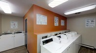 This shared laundry facility is designed with functionality and user convenience at its core. The room features a row of commercial-grade top-loading washing machines lined neatly along a bold orange accent wall, creating a visually distinctive and easy-to-navigate space. Matching dryer units are positioned adjacent, providing a seamless workflow for laundry tasks. The vibrant orange wall serves as both a design focal point and a practical backdrop, displaying clearly posted laundry tip guides and instructional signage to assist users with machine operation and best practices. Overhead, bright fluorescent lighting ensures the room is well-lit throughout the day and evening. The flooring consists of durable wood-look planking, offering easy maintenance suited to a high-traffic communal environment. A textured ceiling adds a subtle architectural detail to the otherwise utilitarian space. A secondary access door is visible along the side wall, providing additional entry and exit convenience. Overall, the laundry facility strikes a balance between practical utility and a clean, organized aesthetic, making it a functional and welcoming amenity for residents or guests.