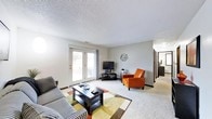 This single-floor residence offers a comfortable and functional living environment centered around a well-proportioned living room. The space is finished with wall-to-wall carpeting that adds warmth underfoot, complemented by neutral gray walls and a textured ceiling that contribute to a clean, understated aesthetic.

The living room serves as the focal point of the home, featuring a set of French doors that flood the space with natural light while providing direct access to an outdoor patio area. Views of the surrounding exterior are visible through the glass panes, offering a pleasant connection to the outdoors. A wall-mounted decorative accent adds a subtle artistic touch to the room.

The layout flows naturally toward a hallway visible at one end of the living room, suggesting additional rooms beyond — likely a bedroom and bathroom. The corridor leads to what appears to be further private spaces, maintaining a clear separation between living and sleeping areas.

Overall, the space presents a straightforward, livable floor plan with adequate natural light, practical finishes, and a cohesive neutral color palette throughout.