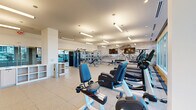 This single-floor fitness center offers a comprehensive workout environment designed to accommodate a wide range of exercise needs. The space is defined by its modern aesthetic, combining clean white walls and ceilings with a striking exposed brick accent wall adorned with a large graphic mural, adding a dynamic visual element to the room. Large floor-to-ceiling windows line one side of the facility, flooding the interior with natural light and offering open views of the surrounding urban landscape, creating an energizing atmosphere for exercise.

The flooring throughout consists of large-format light tile and rubber mat sections, providing both durability and comfort underfoot. Cardio equipment is prominently featured, with a row of treadmills and elliptical machines positioned directly in front of the expansive windows. Stationary recumbent and upright bikes are also available for low-impact training.

The strength training zone is well-appointed with a multi-station cable machine, Smith machines, a variety of free weights, barbells, kettlebells, medicine balls, and resistance bands organized on wall-mounted racks. Full-length mirrors are installed throughout, enhancing the sense of space while supporting proper form during workouts. Pendant and linear LED lighting fixtures complement the recessed ceiling lights, ensuring the space is thoroughly and evenly illuminated at all times.