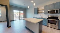 Located at 1648 Eighth Ave in Greeley, Colorado, this single-floor unit presents a thoughtfully designed open layout with contemporary finishes throughout. The kitchen serves as the centerpiece of the space, showcasing blue-grey cabinetry paired with crisp white subway tile backsplash, open wood shelving, and a full suite of stainless steel appliances including a refrigerator, range, microwave, and dishwasher. A central island with a clean white countertop provides additional prep space and is highlighted by elegant pendant lighting overhead. Light wood-tone flooring extends throughout the unit, complementing the cool-toned color palette and teal baseboards that create a cohesive design aesthetic. The living area receives generous natural light through a large sliding glass door that opens to a private balcony with views of the surrounding neighborhood. A separate bedroom features its own window with views of the street, allowing additional natural light to fill the room. The bathroom is equipped with open shelving for storage, a vanity with matching cabinetry, and accessibility-friendly grab bars. Tall ceilings enhance the overall sense of openness and airiness throughout the unit. The space combines modern design elements with practical functionality in a fresh, welcoming atmosphere.