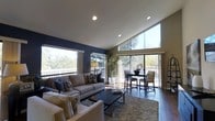 This single-story clubhouse offers a welcoming and well-appointed interior designed for both comfort and functionality. The main living area is defined by soaring vaulted ceilings with recessed lighting, creating a sense of openness and volume. A striking wall of floor-to-ceiling windows and sliding glass doors lines the perimeter, flooding the space with abundant natural light and providing expansive views of the surrounding mature tree canopy. The seamless connection to the outdoor deck enhances the indoor-outdoor flow, making the space ideal for gatherings and relaxation.

The interior features rich hardwood flooring throughout, anchored by a plush area rug that defines the seating area. A generously sized neutral sofa arrangement centers around a sleek, dark coffee table, offering comfortable seating for guests. Adjacent to the lounge area, a bistro-style dining setup with dark wood high-top table and upholstered chairs provides an additional gathering spot. A bold navy accent wall adds a sophisticated contrast to the warm, neutral tones of the furnishings and the light ceiling. The overall ambiance is modern yet inviting, balancing natural elements with contemporary design details.